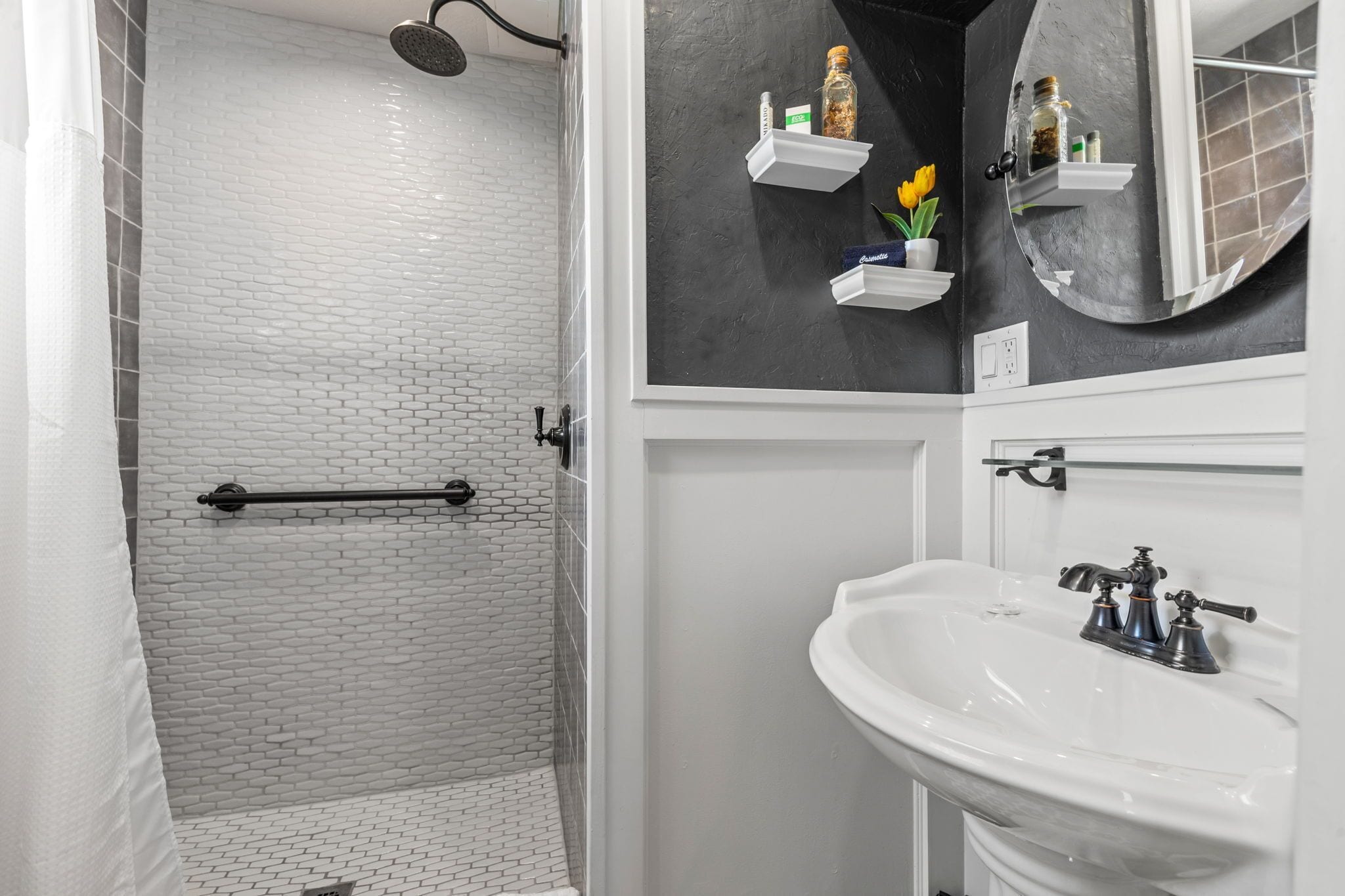 A bathroom with a white brick shower, a white sink with a vintage faucet, and a black wall with shelves holding toiletries and a potted plant.