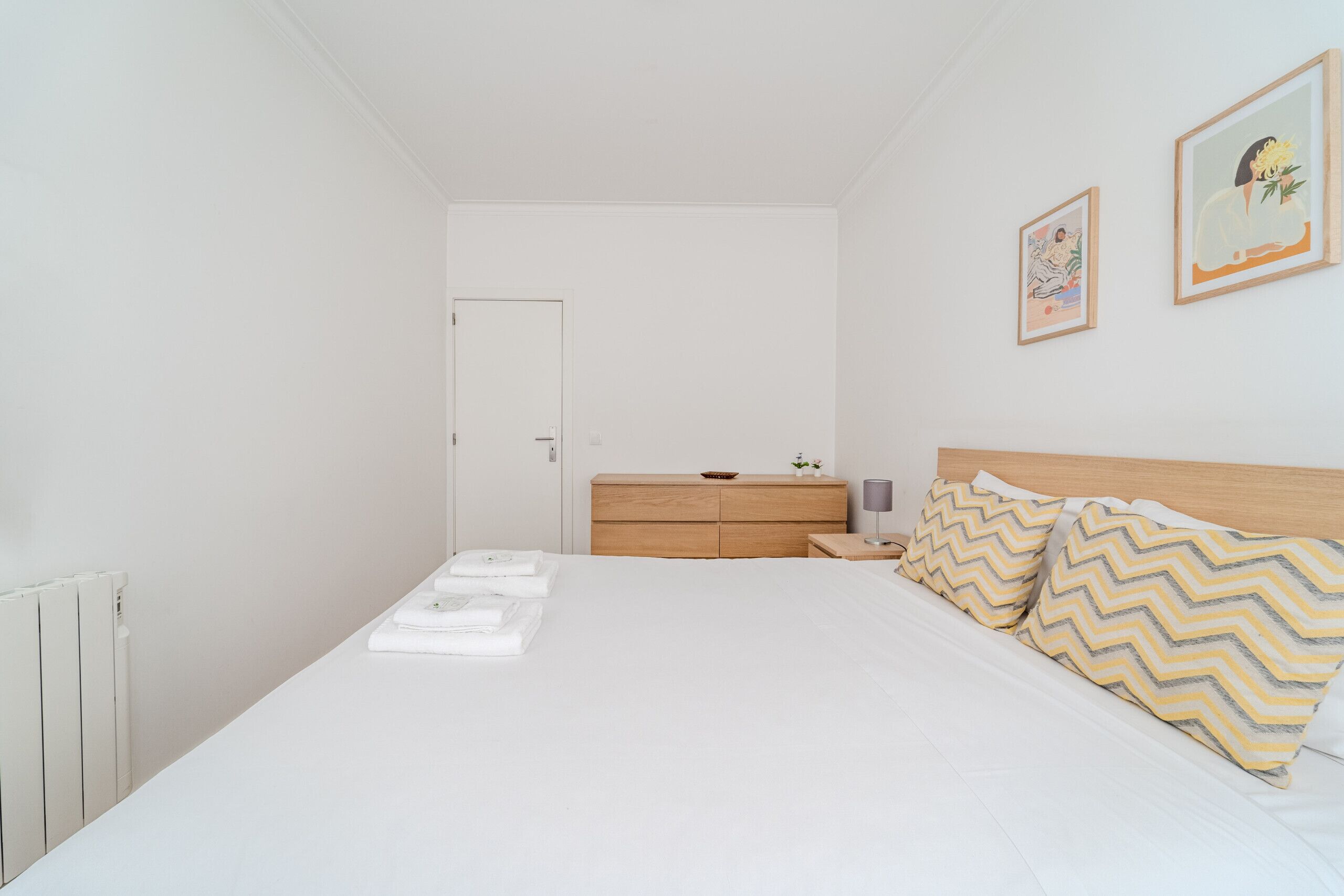 Un dormitorio con una cama, un aparador de madera, una mesita de noche y dos cuadros enmarcados en la pared.