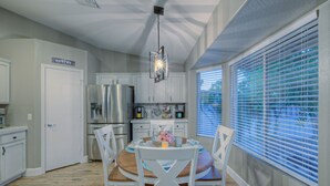 Una cocina moderna con electrodomésticos de acero inoxidable, un comedor con sillas blancas y vistas amplias a través de ventanas con persianas.