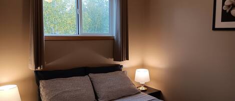 A bedroom with a bed, two bedside lamps, a window with curtains, and a framed picture on the wall.
