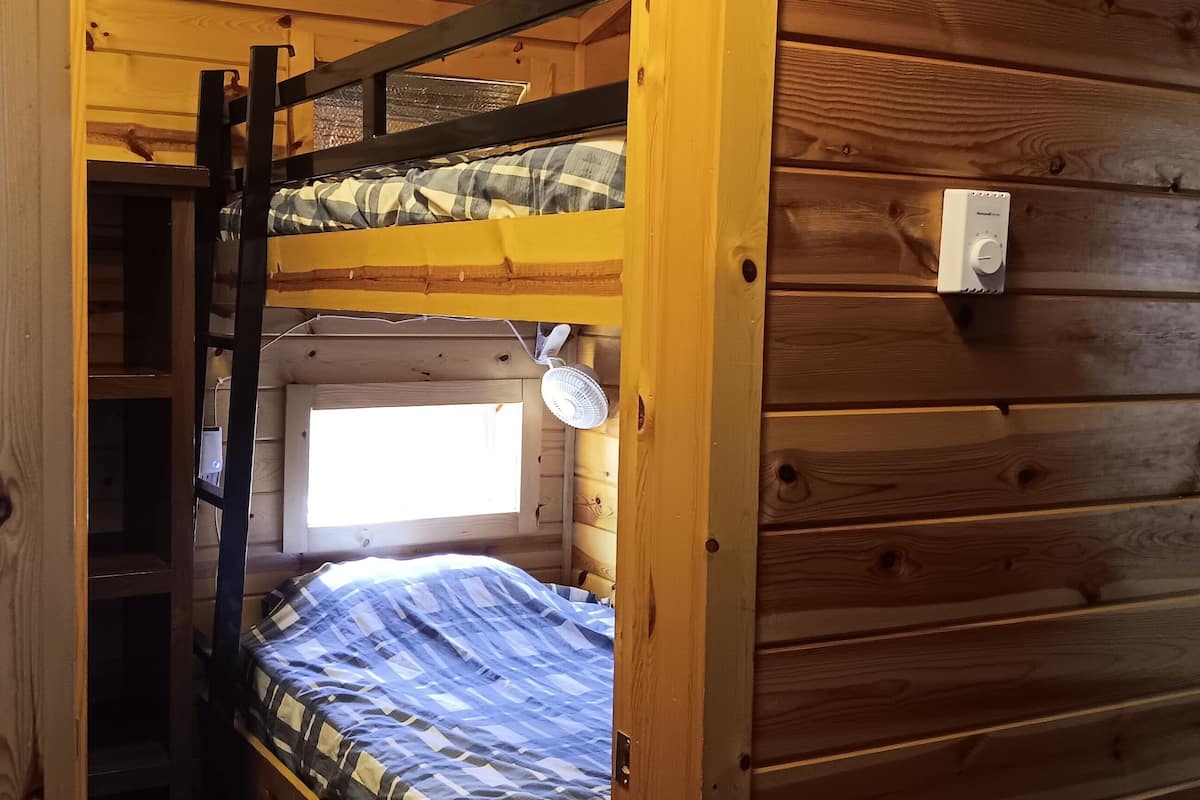 A wooden cabin with a bunk bed, a small window, and a thermostat on the wall.