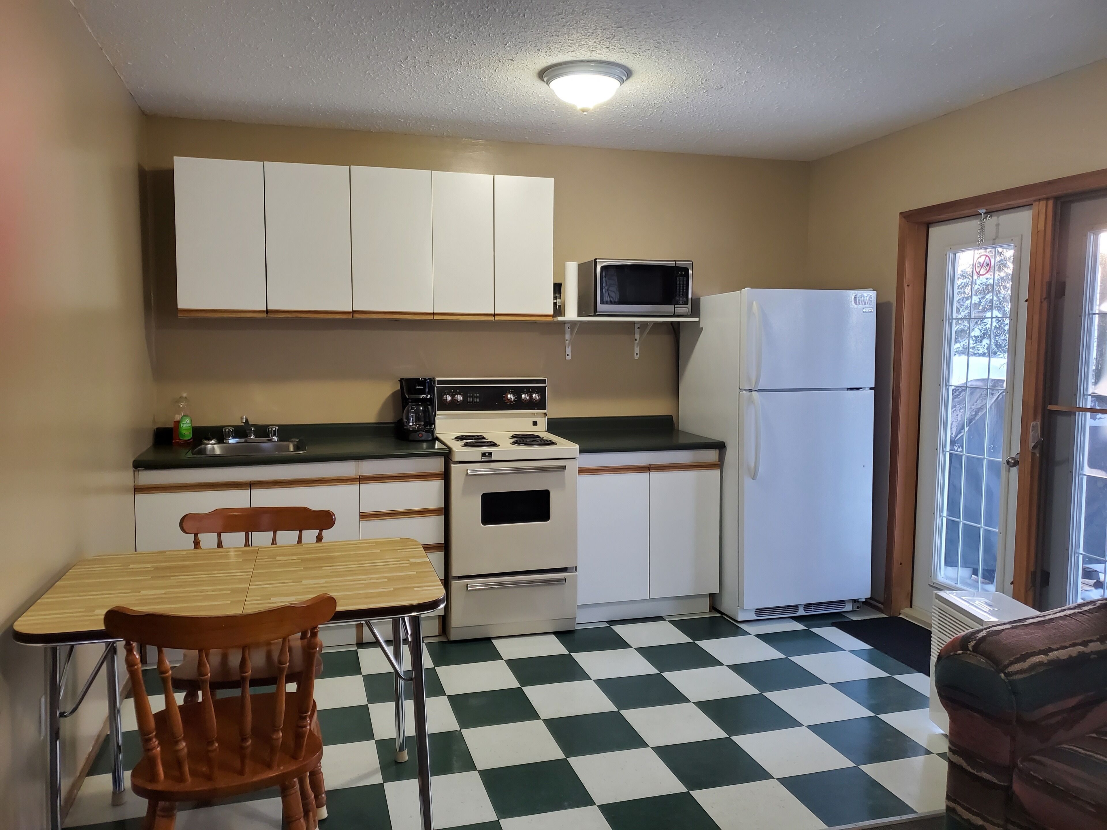 Una cocina funcional con armarios blancos, microondas, estufa y refrigerador. El piso es de cuadros y hay una mesa de madera con sillas.