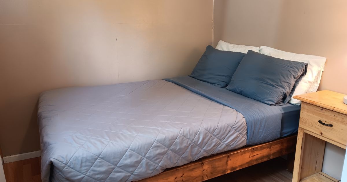 A single bed with a quilted blue mattress and pillows, a wooden headboard, and a bedside table with a drawer.