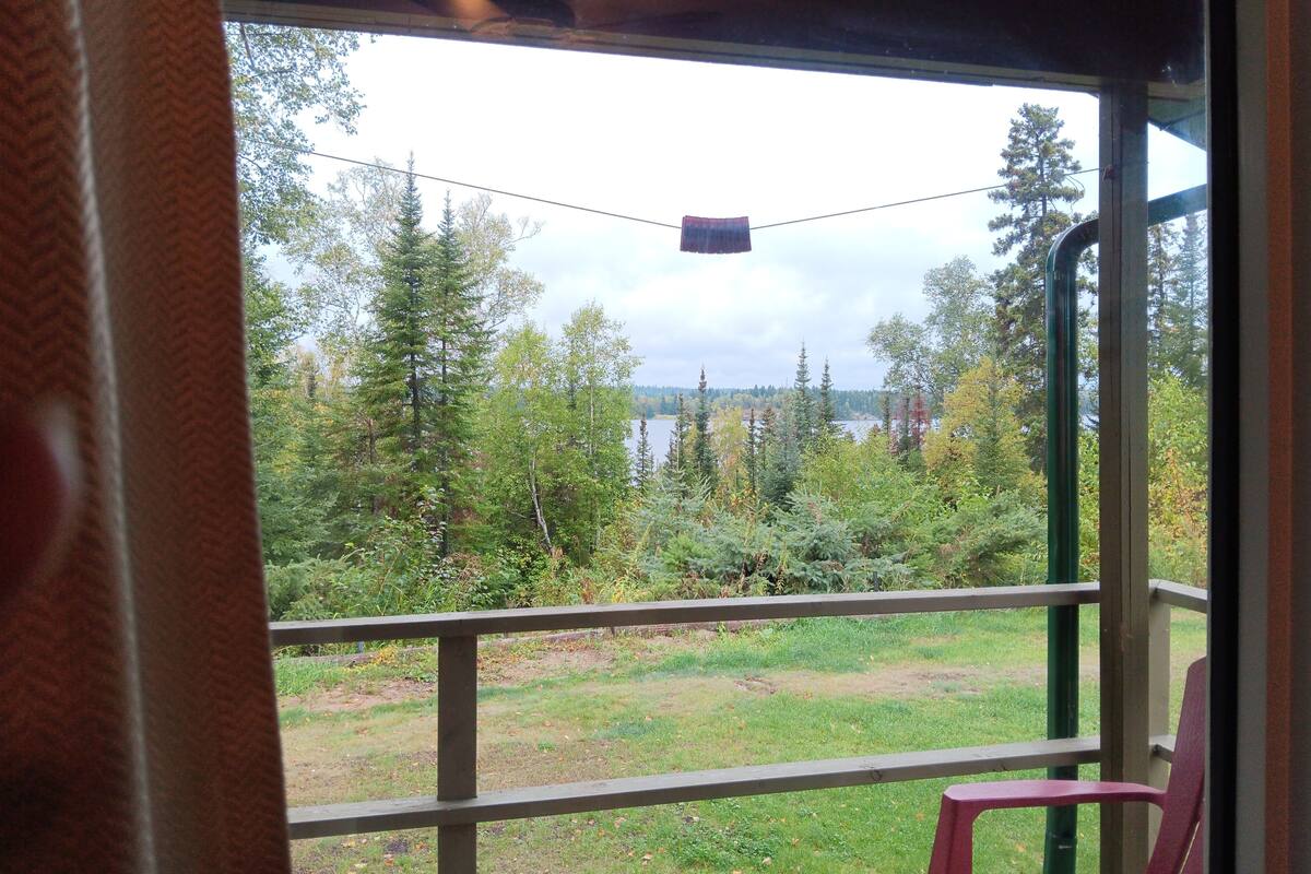 A view from a covered porch with a railing, overlooking a grassy area and dense trees, with a body of water in the background.