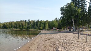 A sandy beach with a calm lake, trees, and playground equipment in the background.