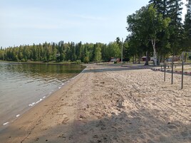 Uma praia arenosa com um lago calmo, árvores e equipamentos de parquinho ao fundo.