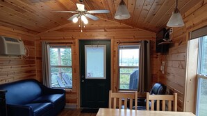 A cozy cabin interior with a wooden ceiling, a ceiling fan, a black door, a dining table with four chairs, a blue leather couch, and two windows with curtains.