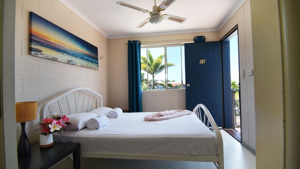 A hotel room with a bed, a nightstand, a painting, a ceiling fan, and a view of palm trees outside. - Caloundra Backpackers - Hostel (Caloundra)