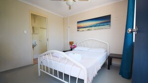 A bedroom with a white metal bed, a painting of a beach scene, a nightstand with a lamp, and a bathroom door. - Caloundra Backpackers - Hostel (Caloundra)