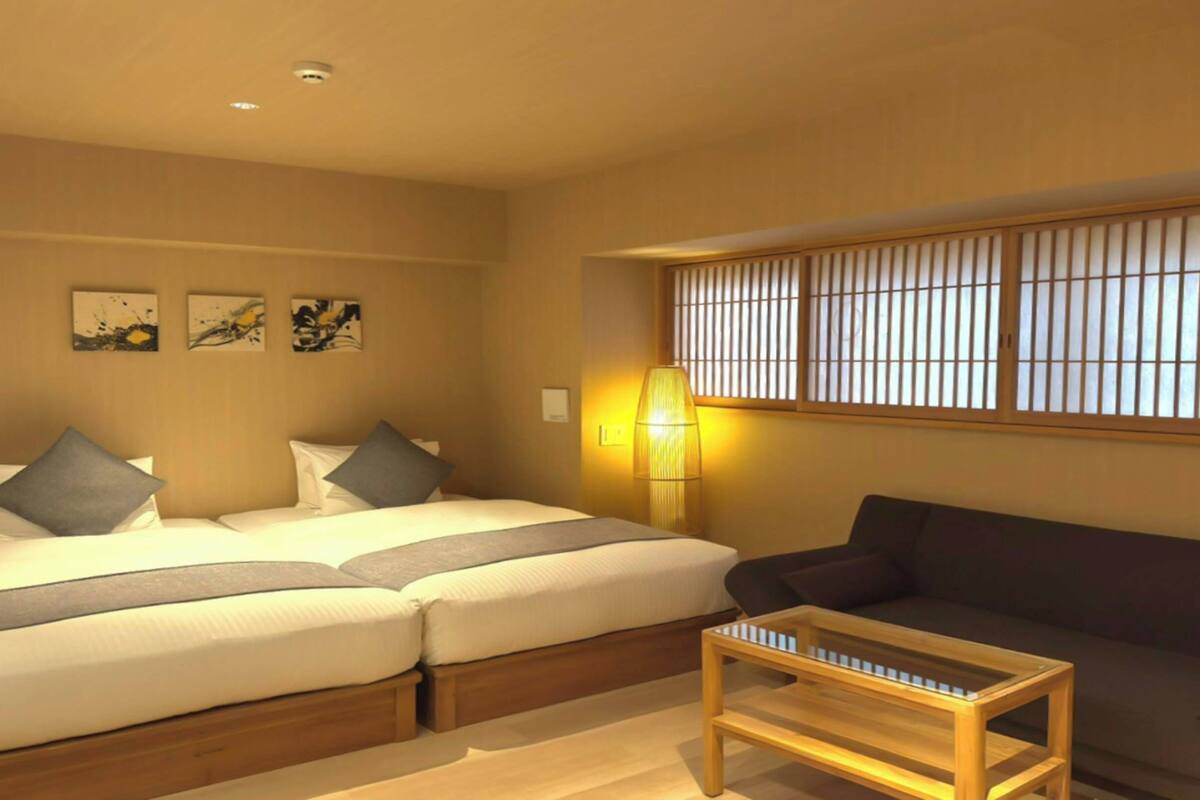A hotel room with two beds, a sofa, a small table, and a window with vertical blinds.
