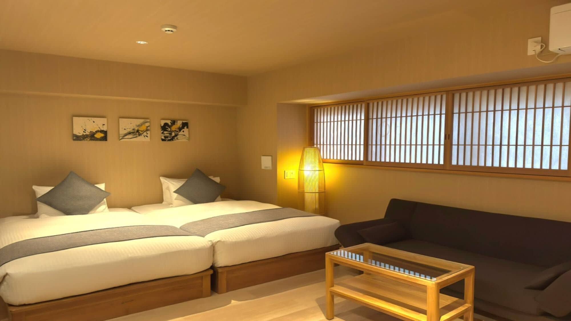 A hotel room with two beds, a sofa, a small table, and a window with vertical blinds.