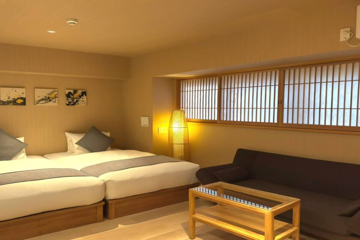 A hotel room with two beds, a sofa, a small table, and a window with vertical blinds.