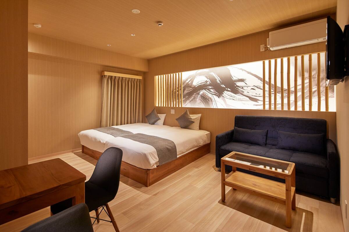 A modern hotel room with a large bed, a dark blue sofa, a wooden coffee table, and a wall-mounted TV.