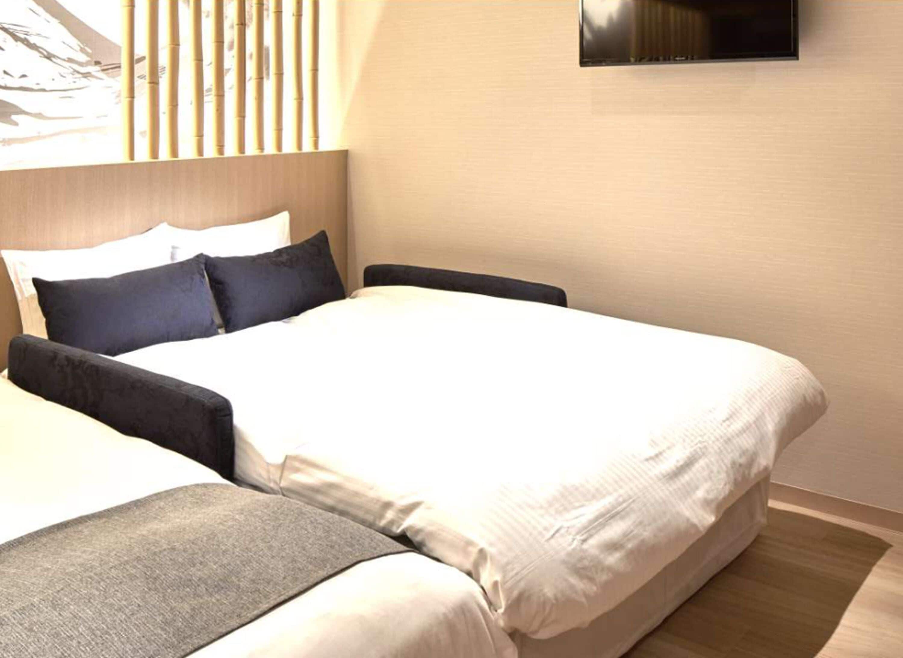 A neatly made bed with white linens and dark pillows, a wooden headboard, and a mounted flat-screen TV on the wall.