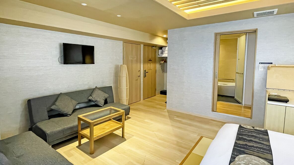 Quarto de hotel moderno com sofá, uma pequena mesa de madeira, televisão de tela plana fixada na parede e guarda-roupa com espelho.