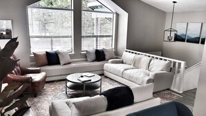 Un salon moderne comprenant un ensemble de canapés, une table basse et un fauteuil. De grandes fenêtres équipées de stores, un luminaire suspendu et un escalier complètent l’ensemble.