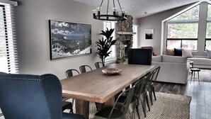 Un espace de repas moderne avec une table en bois, des chaises et un lustre. On aperçoit un canapé et une grande fenêtre à l’arrière-plan.