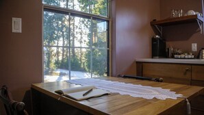 Una mesa de madera con mantel de encaje, un libro y un bolígrafo. Se ve una ventana con vistas a árboles y una puerta con una silla.