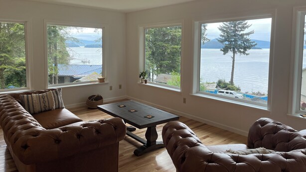 A living room with a large window overlooking a lake, a brown leather sofa, and a dark wooden coffee table. - Waterfront Auke Bay cottage in Juneau (Juneau)