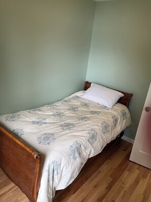 A single bed with a floral-patterned bedspread and a white pillow in a room with light green walls and wooden flooring.