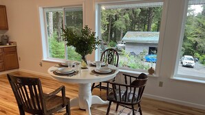A dining area with a round table set for four, surrounded by wooden chairs. Large windows offer a view of a forested area and a road with vehicles. - Waterfront Auke Bay cottage in Juneau (Juneau)