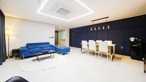 Ein modernes Wohnzimmer mit einem blauen Ecksofa, einem Essbereich mit weißen Stühlen und einer dunkel gestrichenen Akzentwand.