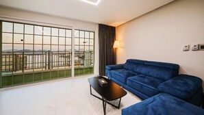 Ein modernes Wohnzimmer mit einem blauen Sofa, einem kleinen schwarzen Tisch und einer großen Schiebeglasdoor, die auf einen Balkon mit Blick auf die Stadt führt.