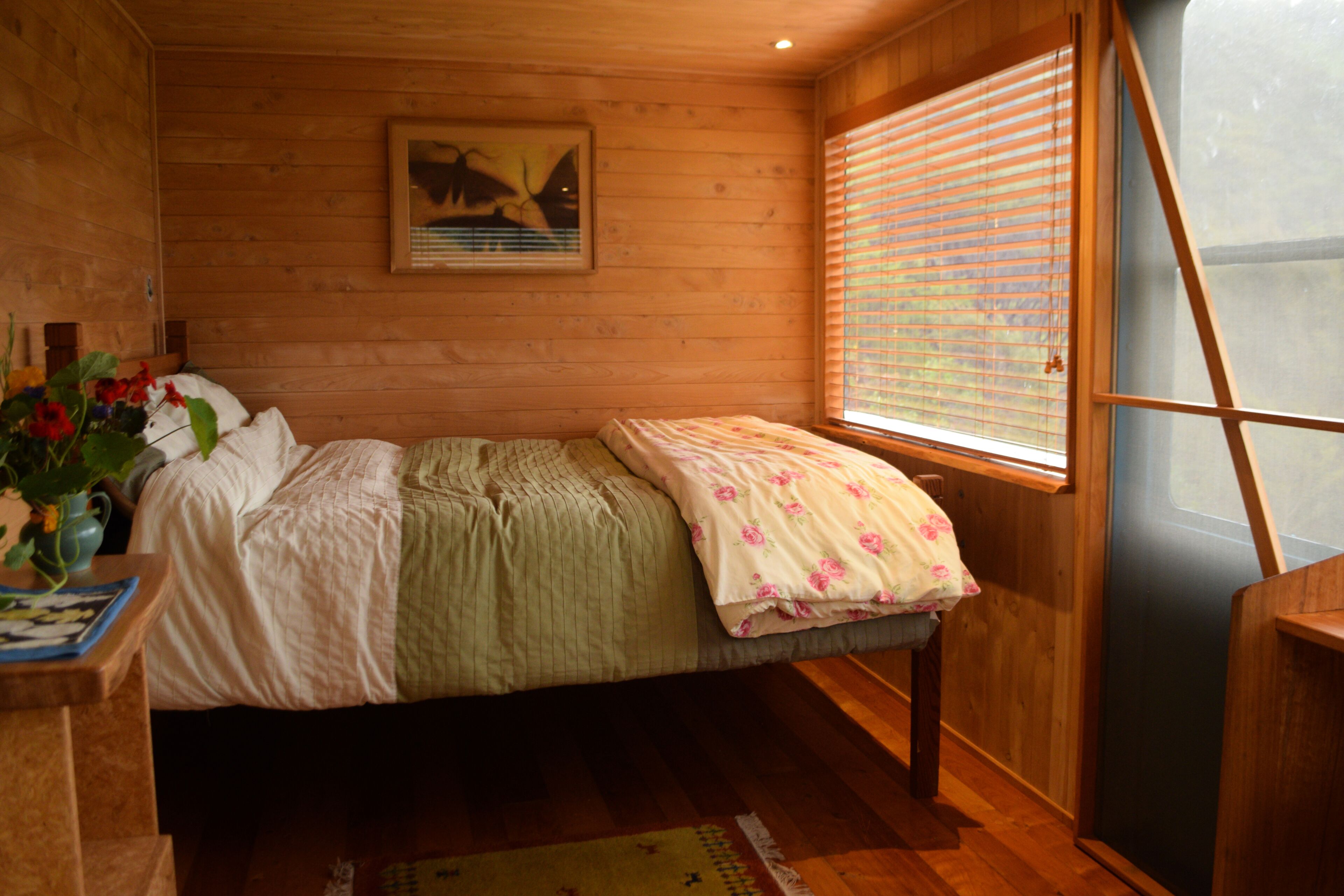A wooden cabin room with a bed, a window with blinds, a painting on the wall, and a small table with a vase of flowers.