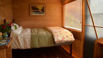 A wooden cabin room with a bed, a window with blinds, a painting on the wall, and a small table with a vase of flowers.