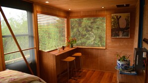 Una habitación cálida tipo cabaña con una cama, un escritorio con dos taburetes, una ventana con persianas, un cuadro y una pequeña mesa con una planta en maceta.
