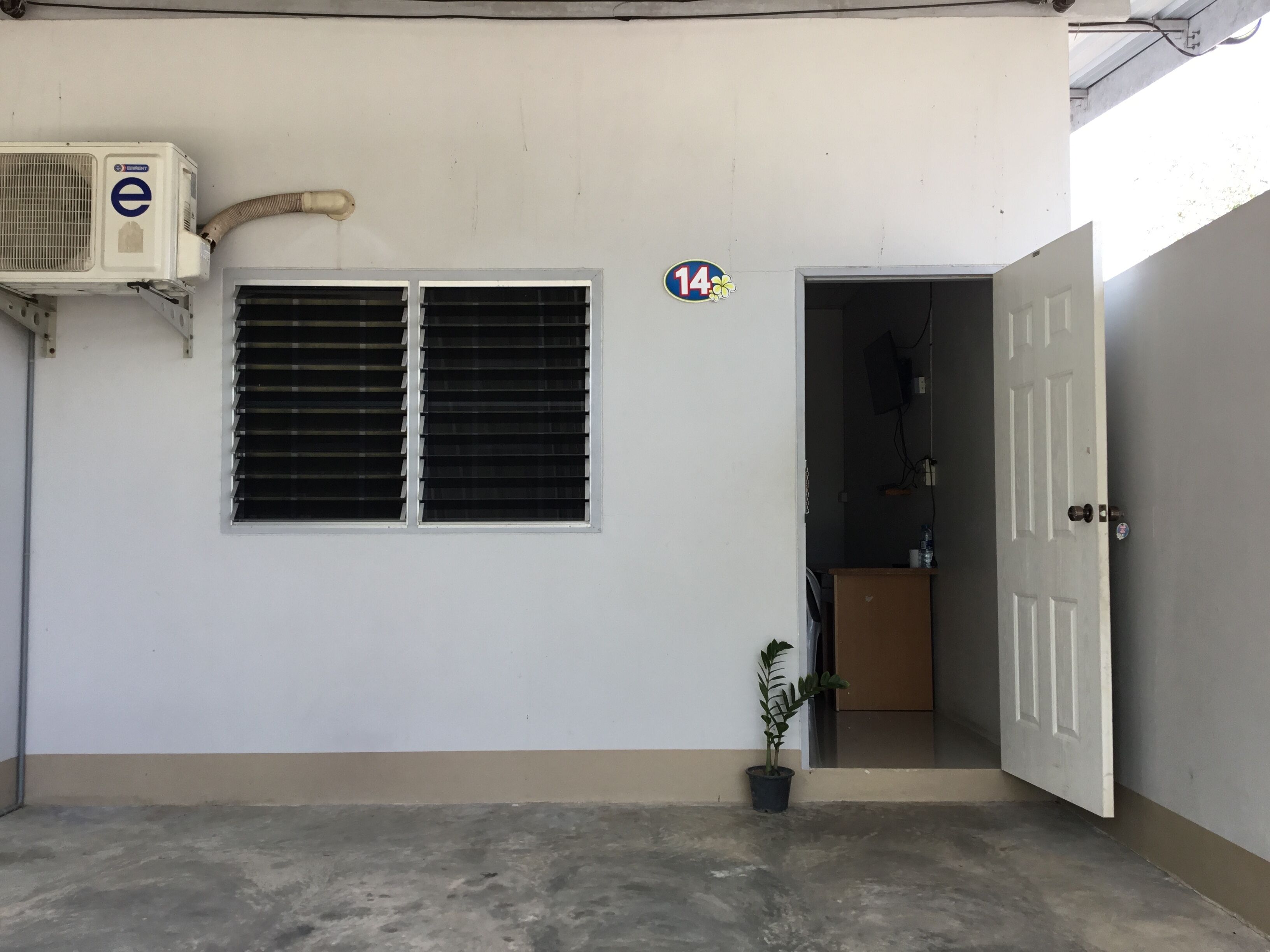 Fachada de un edificio con un muro blanco, dos ventanas con persianas horizontales, una planta en maceta y una puerta.