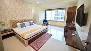 A modern hotel room with a large bed, a desk, a chair, a TV, and a window with a city view. - Blueberry Inn (Raipur)