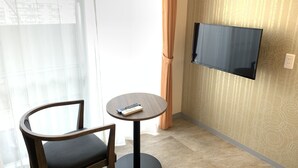 A hotel room with a single round table, a wooden chair, a flat-screen TV mounted on the wall, and a window with sheer curtains. - Mr.KINJO Miki24 in front of Makishi Station (Naha)
