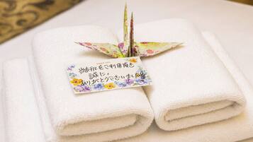 Dos toallas blancas dobladas ordenadamente, una encima de la otra, junto a una grulla de papel decorativa y una tarjeta con texto japonés.