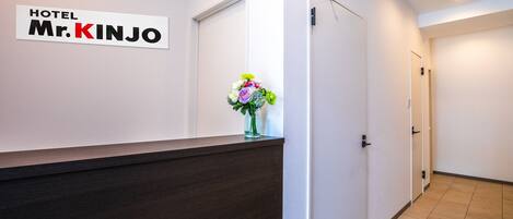Uma área de recepção de hotel com um balcão escuro, uma placa na parede com a inscrição "HOTEL Mr. KINJO" e um vaso com flores.