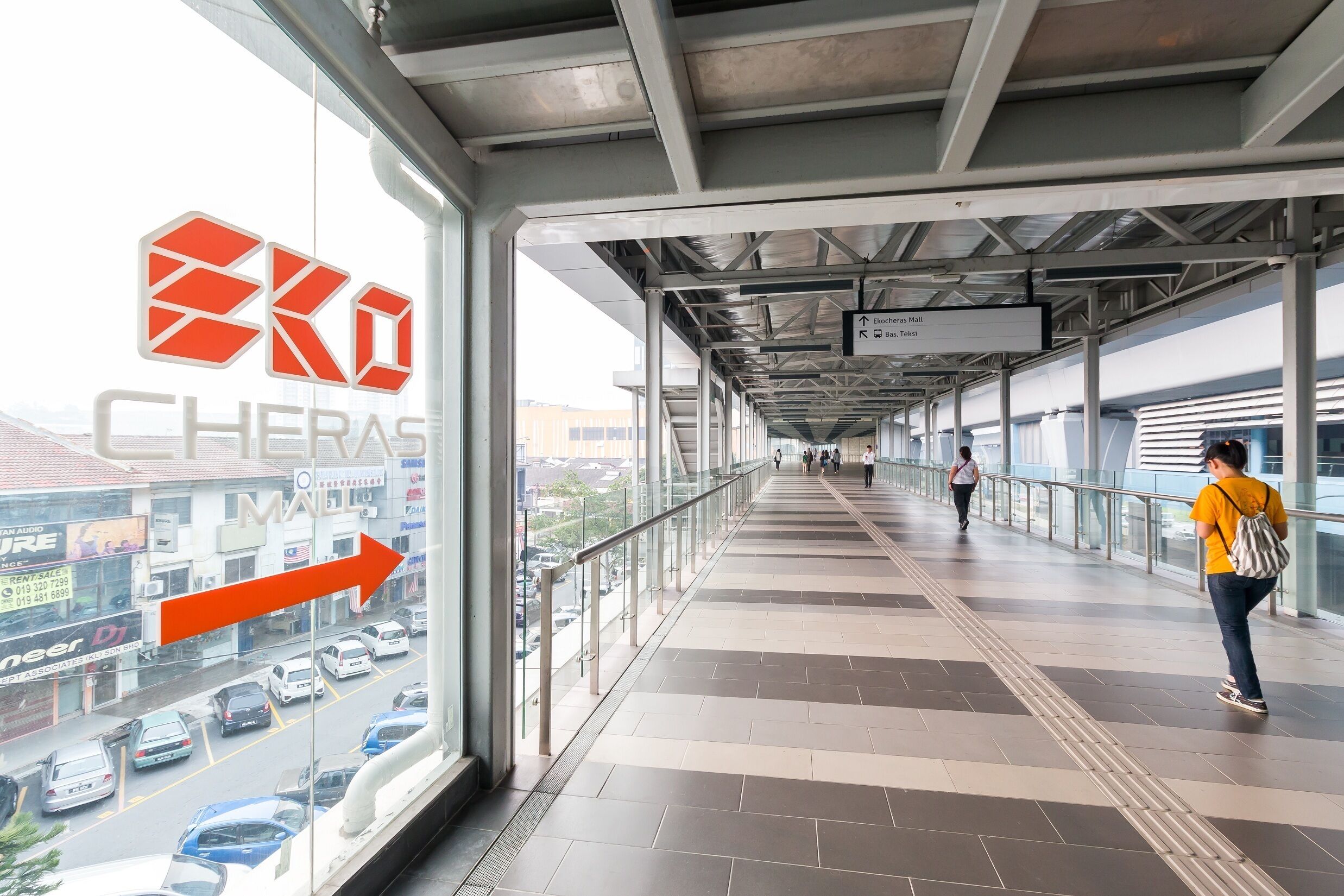 A covered walkway with a view of a busy street, directional signs, and a logo for 'Eko Cheras Mall'.