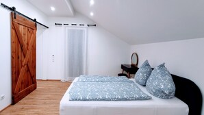 Um quarto com piso de madeira, cama com roupa de cama estampada, porta de celeiro deslizante, uma mesinha e uma cadeira.