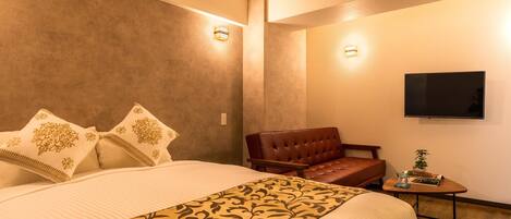 A hotel room with a large bed, a leather armchair, a small table with a plant, and a flat-screen TV mounted on the wall.