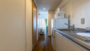 A narrow hallway leading to a bedroom with a bed and a window with green curtains. A kitchenette with a refrigerator, microwave, and sink is visible. - Mr.KINJO in front of Manza Beach (Onna)