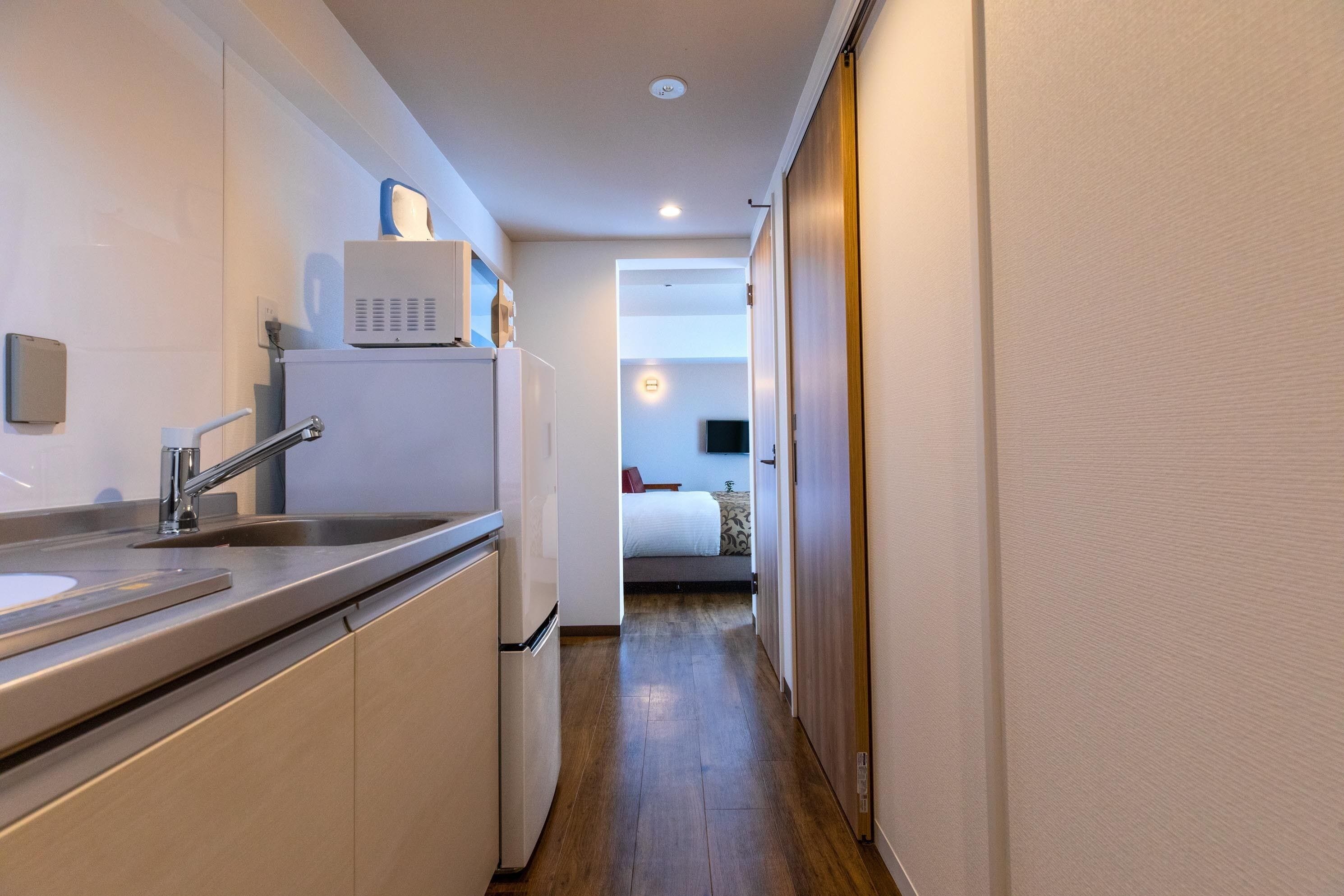 Un pasillo estrecho que conduce a un dormitorio con cama, una pequeña kitchenette con pileta y microondas, y piso de madera.