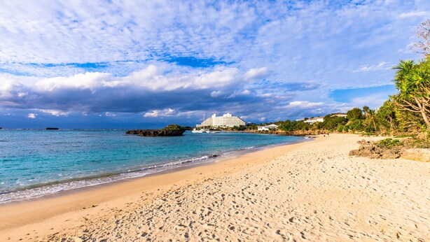 A beach with clear turquoise water, a sandy shore, and a large white building in the background. - Mr.KINJO in front of Manza Beach (Onna)