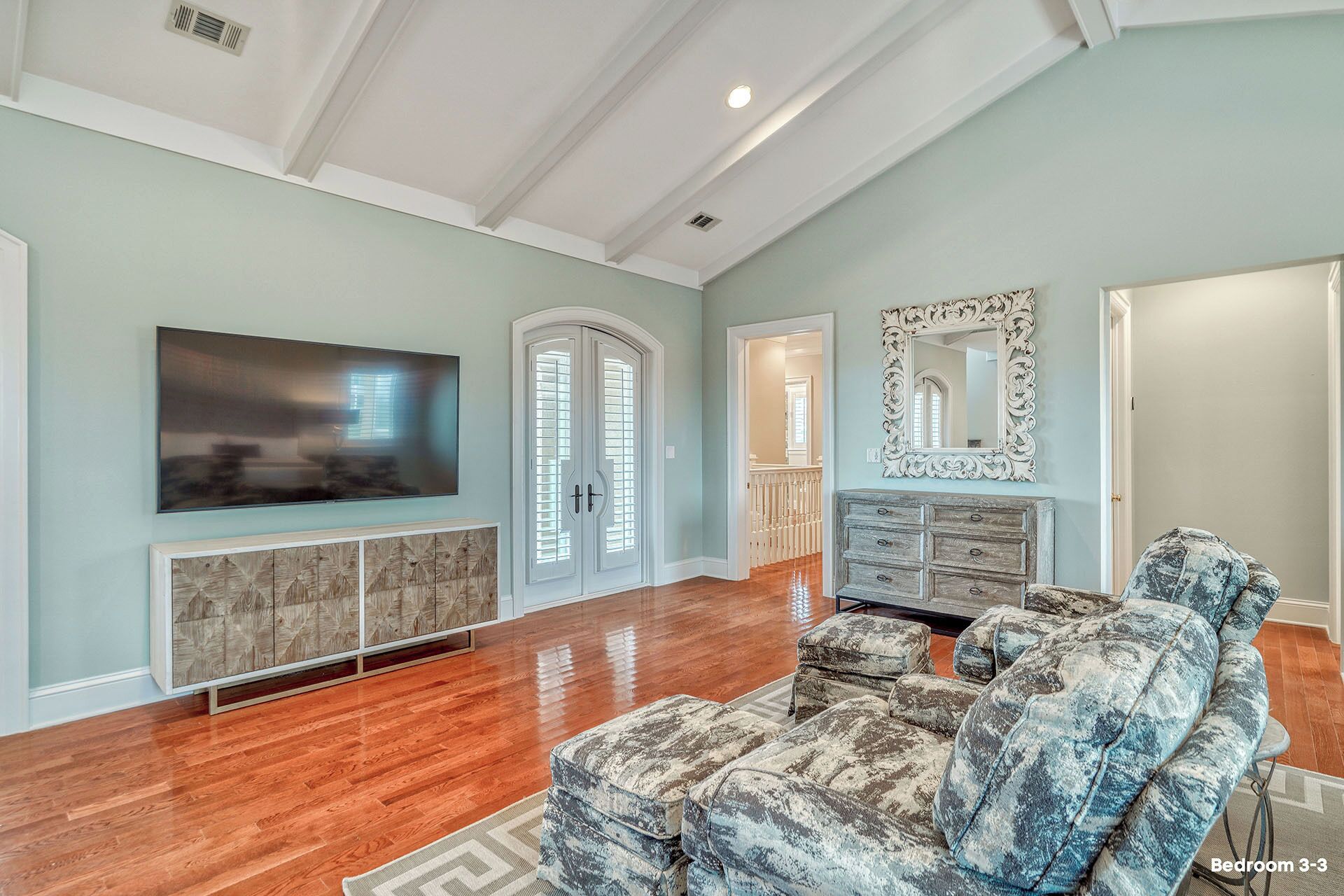 A spacious living room with hardwood flooring, a large flat-screen TV, a built-in entertainment unit, a patterned sofa, and a decorative mirror.