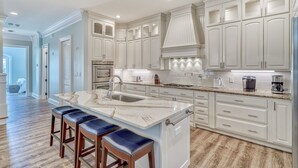 Una cocina moderna con armarios blancos, una isla de mármol y horno empotrado. Hay tres taburetes de bar y una cafetera en la encimera.
