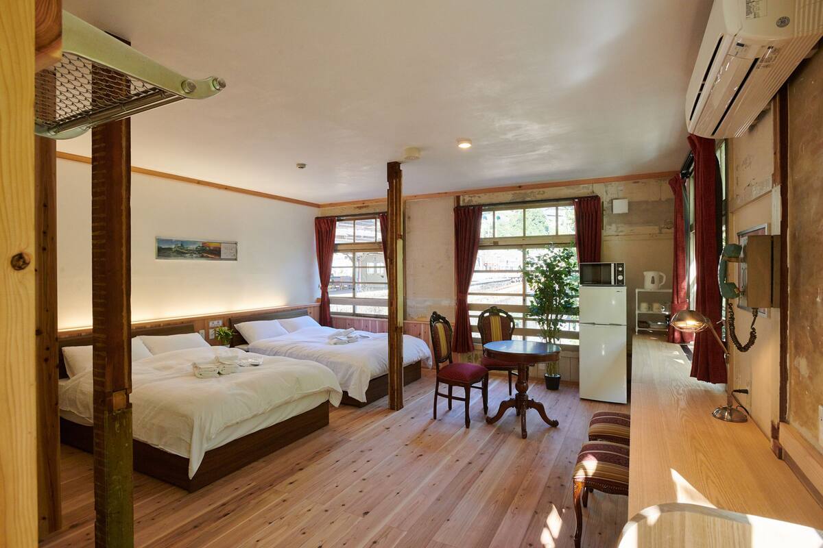 A room with two beds, a dining table, a chair, and a kitchenette.