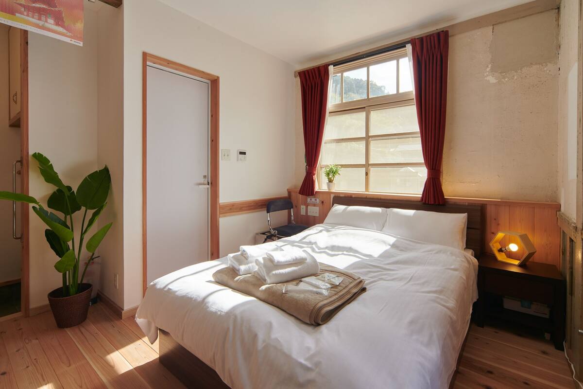 A neatly made bed with white linens, a bedside table with a lamp, a potted plant, and a window with curtains.