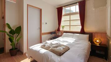 Una cama bien hecha con ropa blanca, una mesita de noche con lámpara, una planta en maceta y una ventana con cortinas.