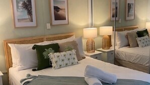 Una cama bien tendida con una manta verde, una toalla blanca y un cabecero de madera. Hay dos mesitas de noche con lámparas, y un espejo grande en la pared.