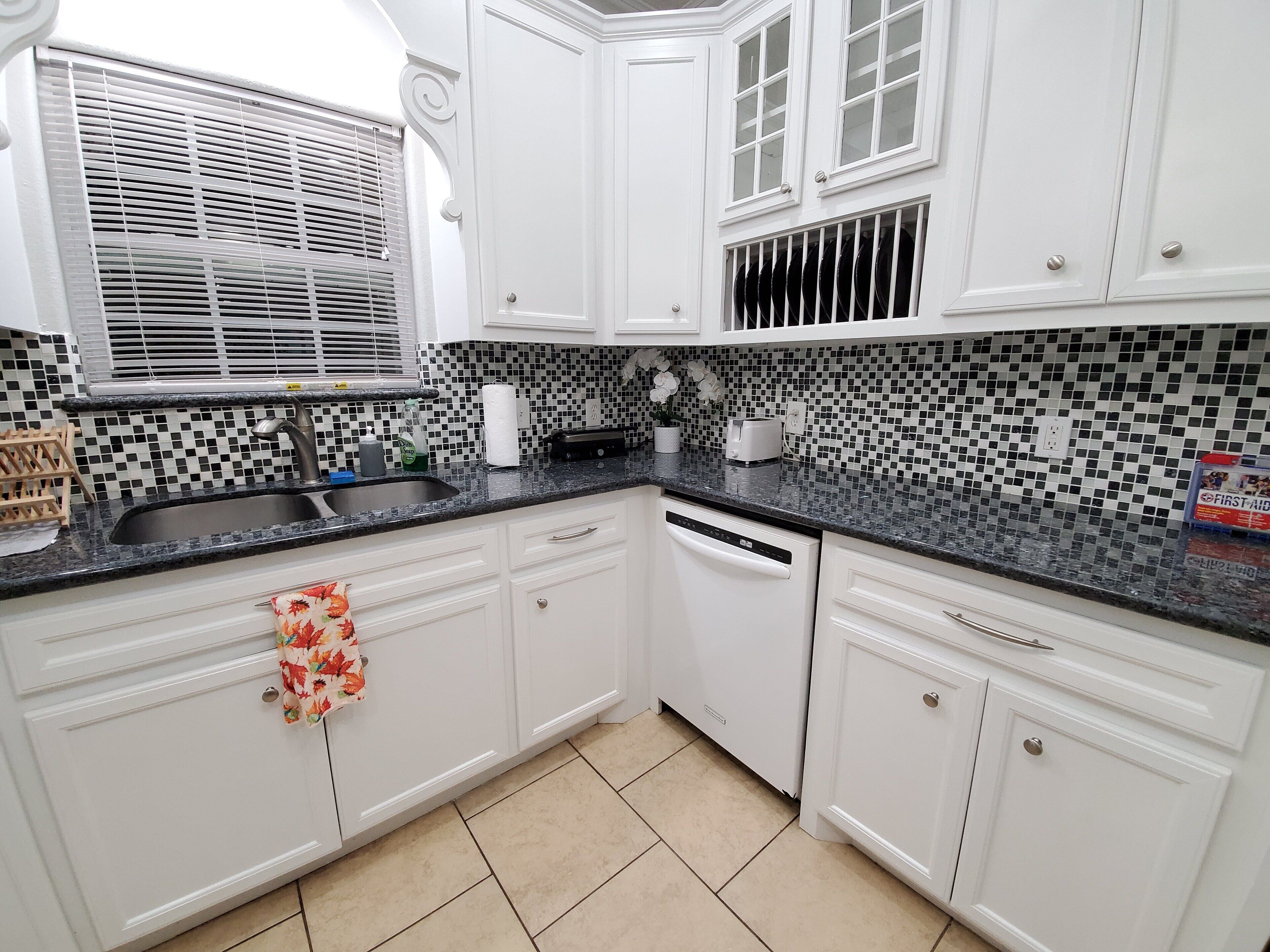 A kitchen with white cabinets, a black countertop, a dishwasher, and a window with blinds.
