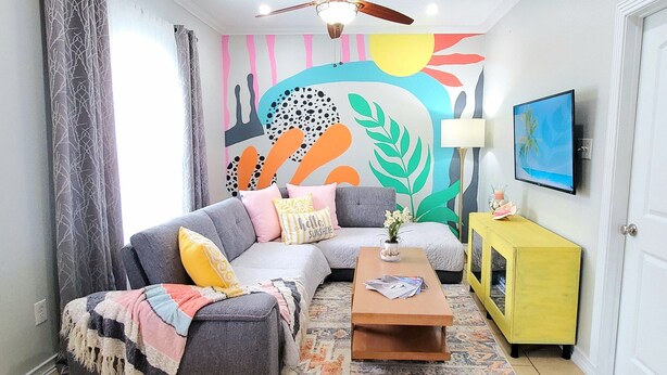 Un salon moderne avec un canapé d’angle, une table basse, un buffet jaune et une fresque murale abstraite et colorée.