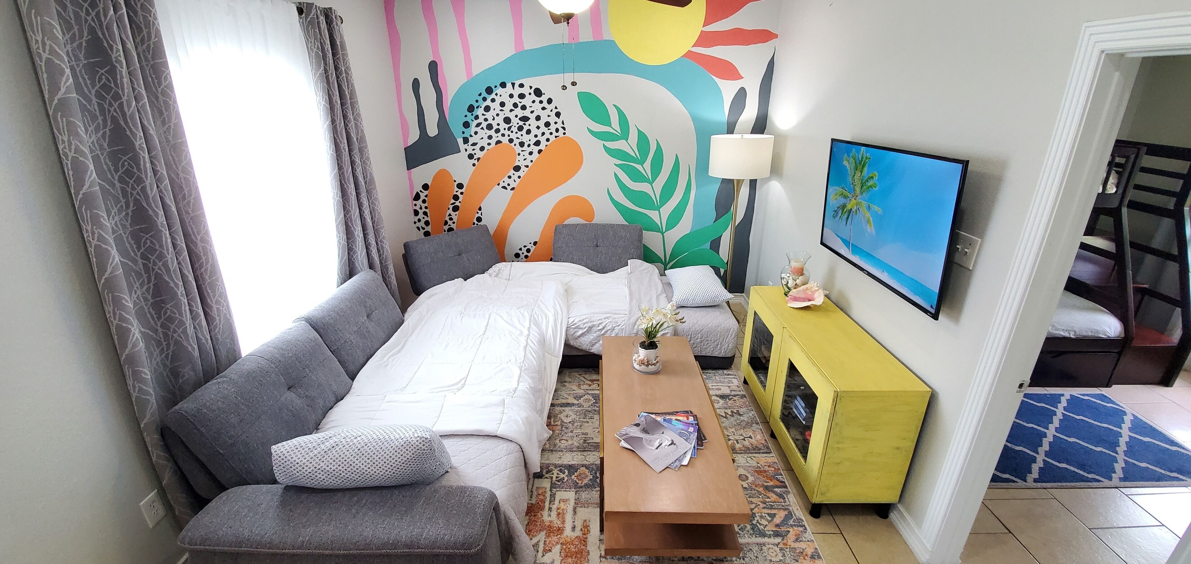 Una habitación acogedora con sofá, una mesita, un mural de pared colorido, una lámpara y televisor.